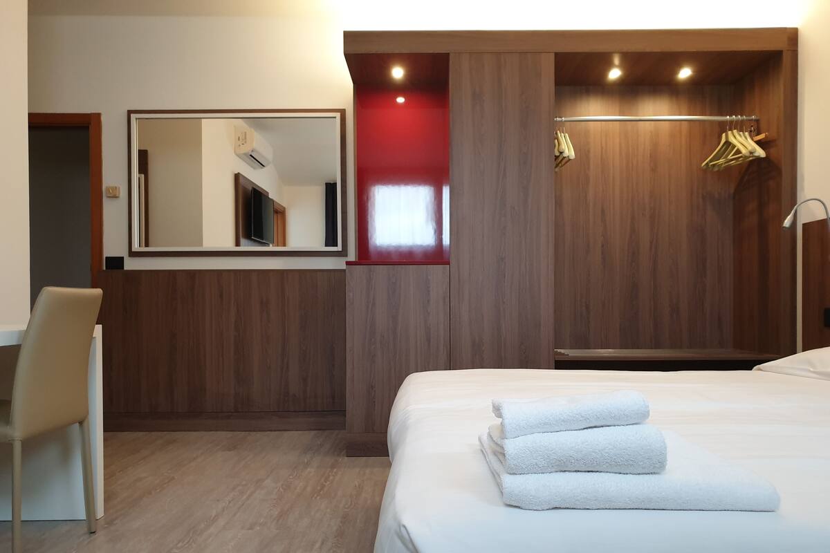 A hotel room with a bed, a desk, a chair, a mirror, and a wardrobe with hanging clothes.