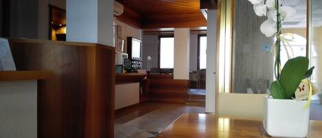 Un espace d’accueil avec un comptoir de réception en bois, un vase blanc orné d’une orchidée et une vue sur la salle à manger.