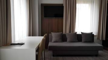 A modern hotel room with a white desk, a grey sofa with cushions, a TV mounted on the wall, and large windows with curtains.