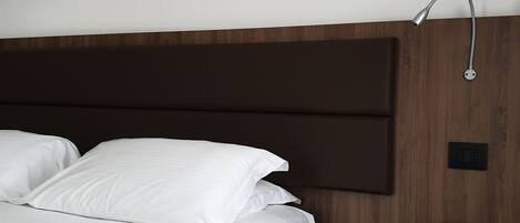Una cama bien hecha con sábanas blancas, cabecera de madera y un cuadro colgado en la pared que muestra un paisaje de montaña.