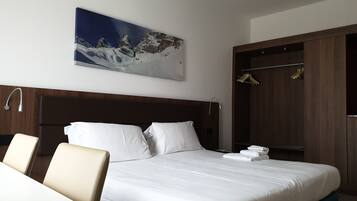 Ein Hotelzimmer mit einem großen Bett, einem Holzkopfende, einem Gemälde einer Berglandschaft, einem Kleiderschrank und einer Sitzecke mit Tisch und Stühlen.