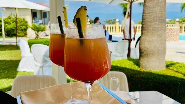 Two glasses of a red beverage with straws and a pineapple slice on a table outdoors with umbrellas and palm trees in the background. - Grand Hotel Stella Maris Italia (Palmi)