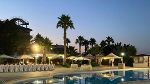 A pool area with tables and chairs, palm trees, and a building in the background. - Grand Hotel Stella Maris Italia (Palmi)
