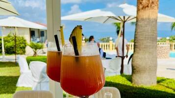 Due bicchieri di una bevanda rossa con cannucce e una fetta di ananas su un tavolo all'aperto, con ombrelloni e palme sullo sfondo.