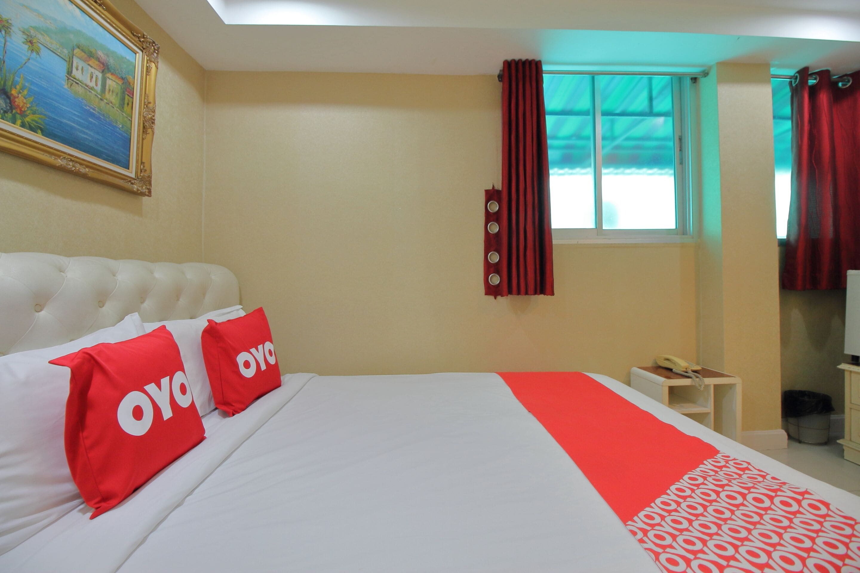 A hotel room with a large bed, two red pillows with 'OYO' branding, a red and white patterned bedspread, a painting on the wall, a window with red curtains, and a small bedside table with a telephone.