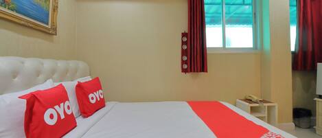 A hotel room with a large bed, two red pillows with 'OYO' branding, a red and white patterned bedspread, a painting on the wall, a window with red curtains, and a small bedside table with a telephone.