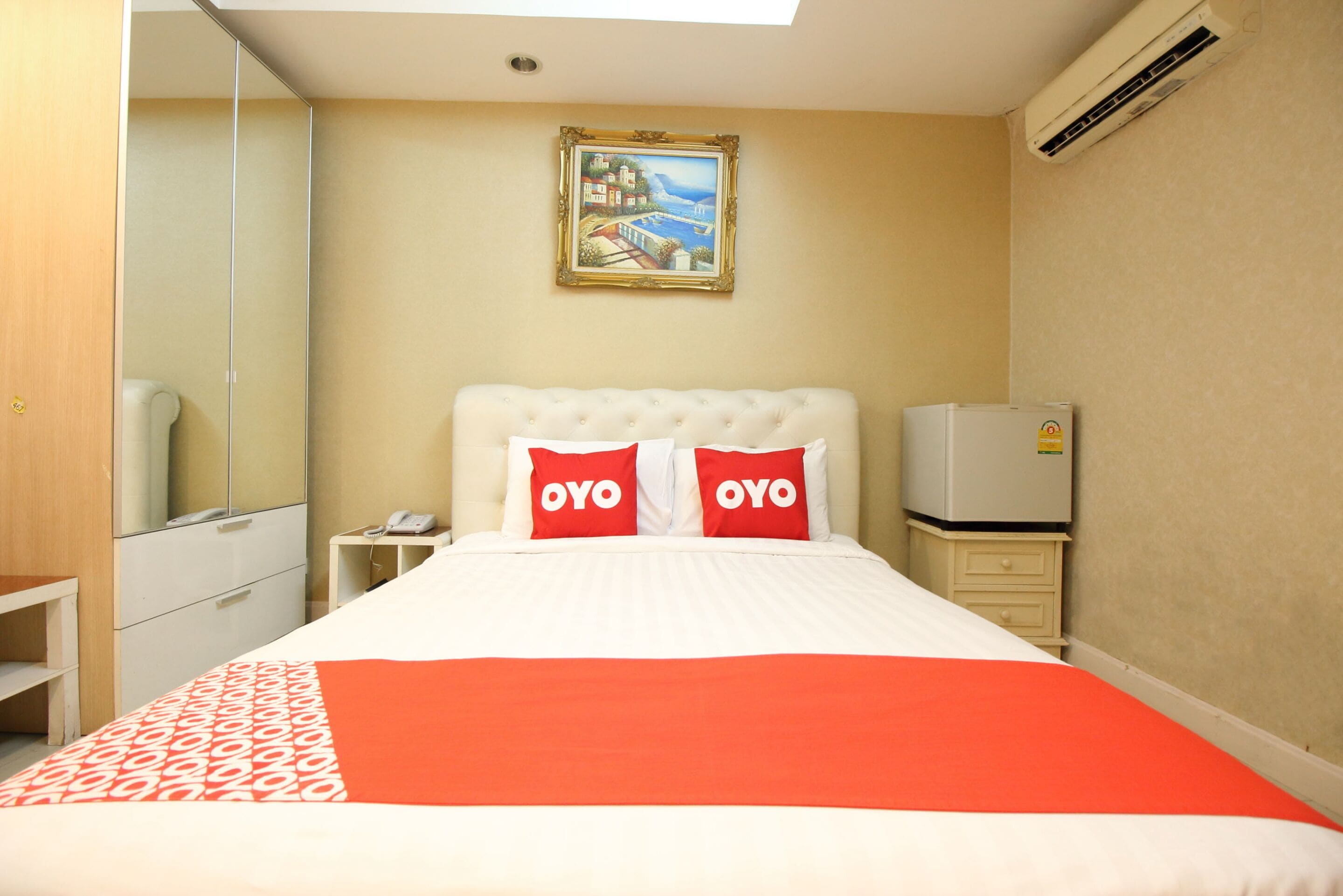 Hotelkamer met een bed, twee kussens met "OYO" branding, een nachtkastje met telefoon en een kleine koelkast, een andere nachtkastje, een spiegel en een schilderij aan de muur.