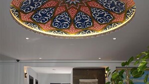 Un hall de hotel con un gran diseño de techo ornamentado, piso de mármol pulido y una recepción con una planta y un cuadro.