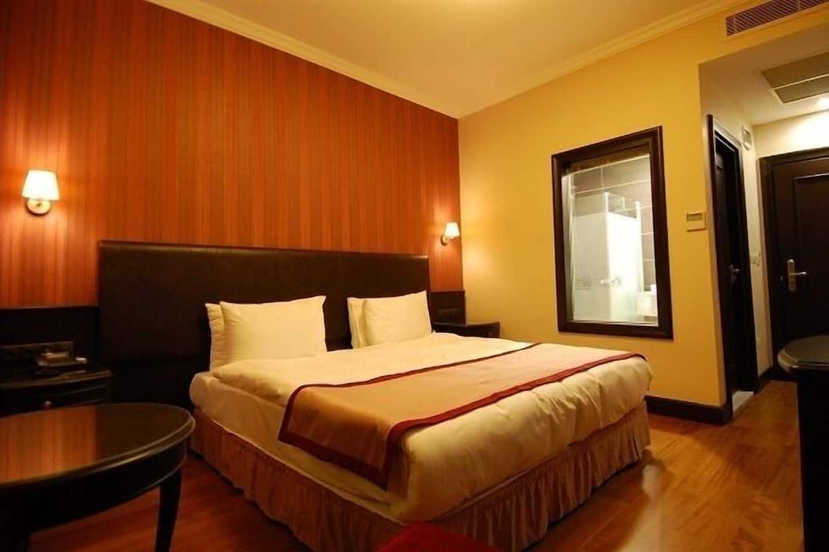 A hotel room with a large bed, wooden panel walls, two bedside lamps, a small table, and a window with a view.