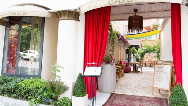 Exterior de un hotel con zona de café, con cortinas rojas, mesas al aire libre y un cartel que enumera los servicios del café.