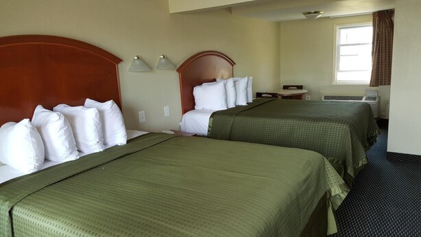 A hotel room with two beds, each with green bedding and white pillows, a wooden headboard, a small table, and a window with curtains. - Royal Inn (Tonawanda)