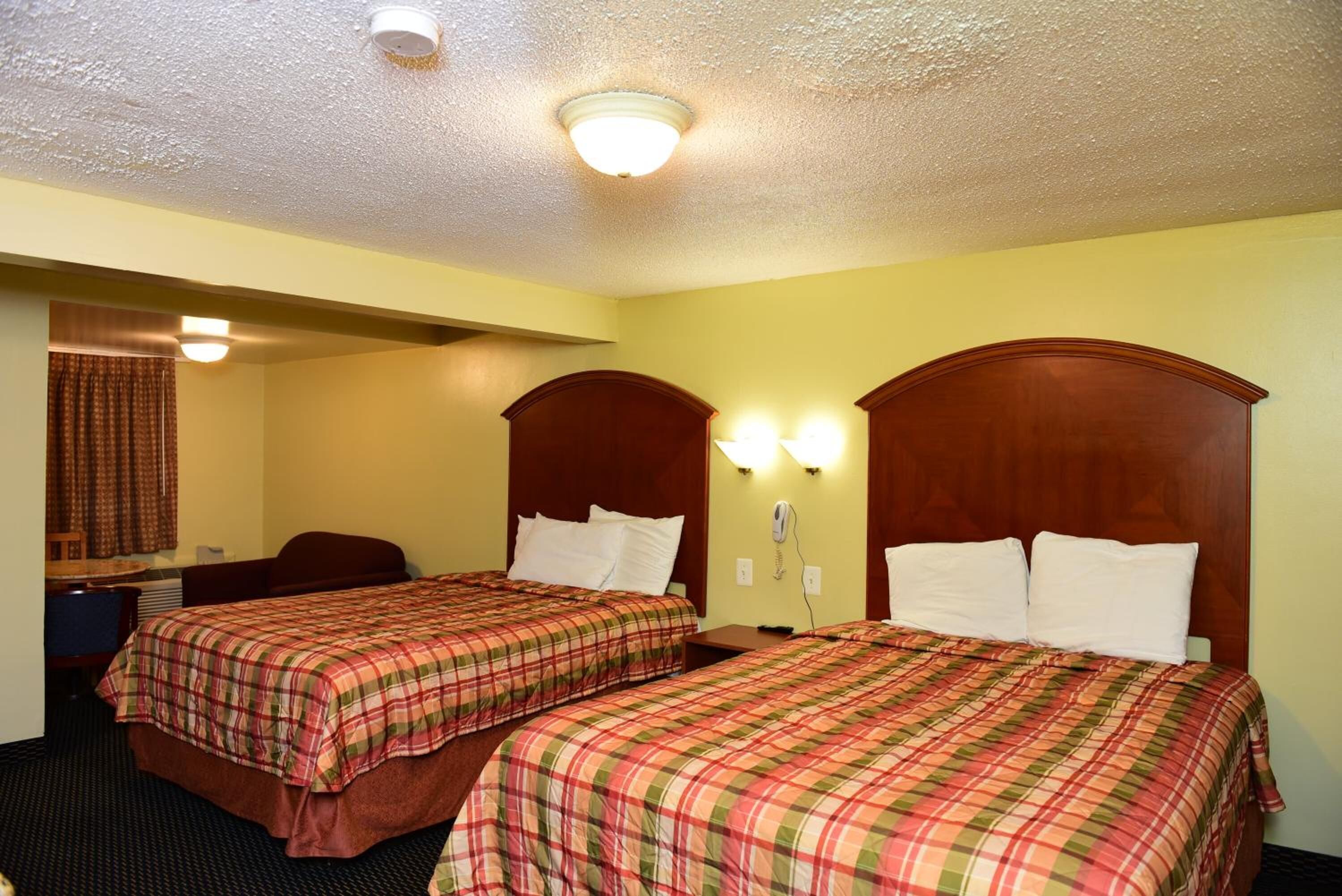 A hotel room with two beds, each with a wooden headboard and a plaid bedspread. There is a desk with a chair and a telephone on the wall.
