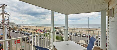 A balcony with a table and chairs, overlooking a beach and a busy street.