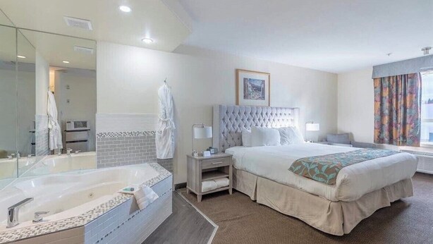A hotel room with a large bed, a separate bathroom with a bathtub, a bedside table with a lamp, and a window with curtains. - Foxwood Inn & Suites (Drayton Valley)
