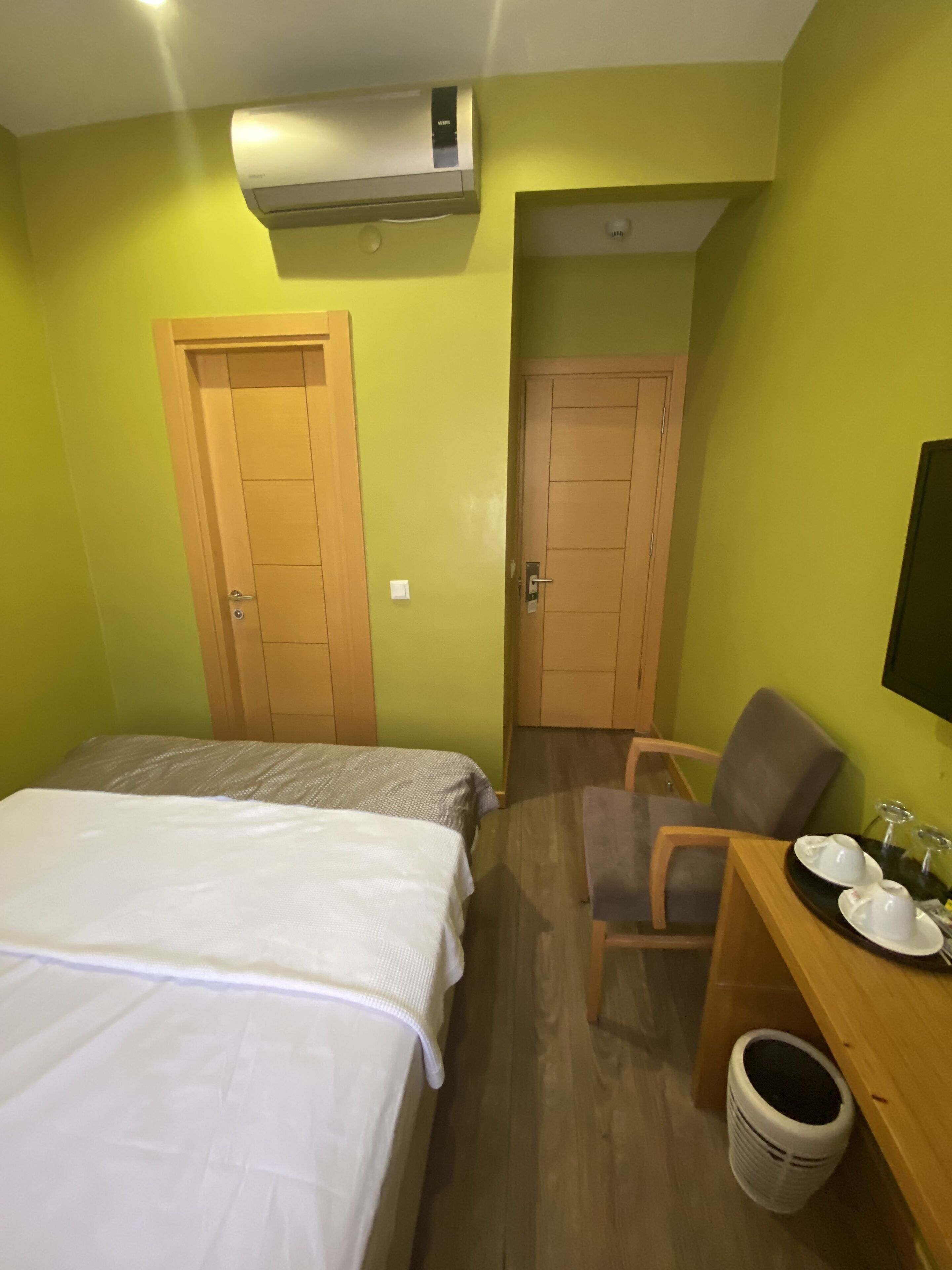 A hotel room with two beds, a desk with a chair, a small table with a tray, and an air conditioning unit.