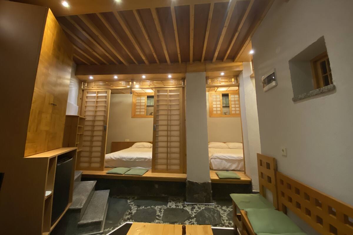 A room with a wooden ceiling, two beds with green pillows, a small table, and a stone-patterned floor.