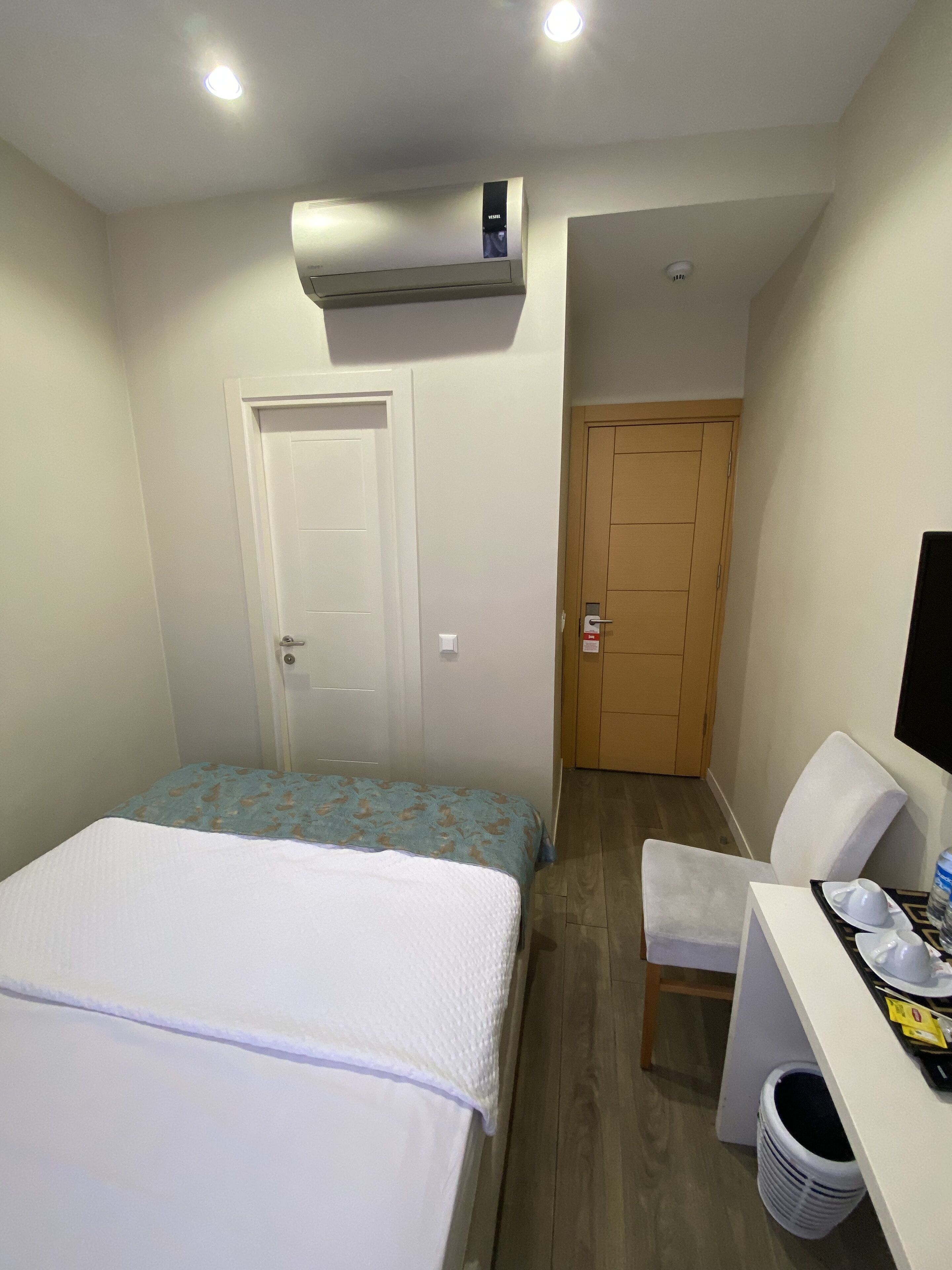 A compact hotel room with a single bed, a desk with a chair, a small table with a water bottle, and a trash bin.