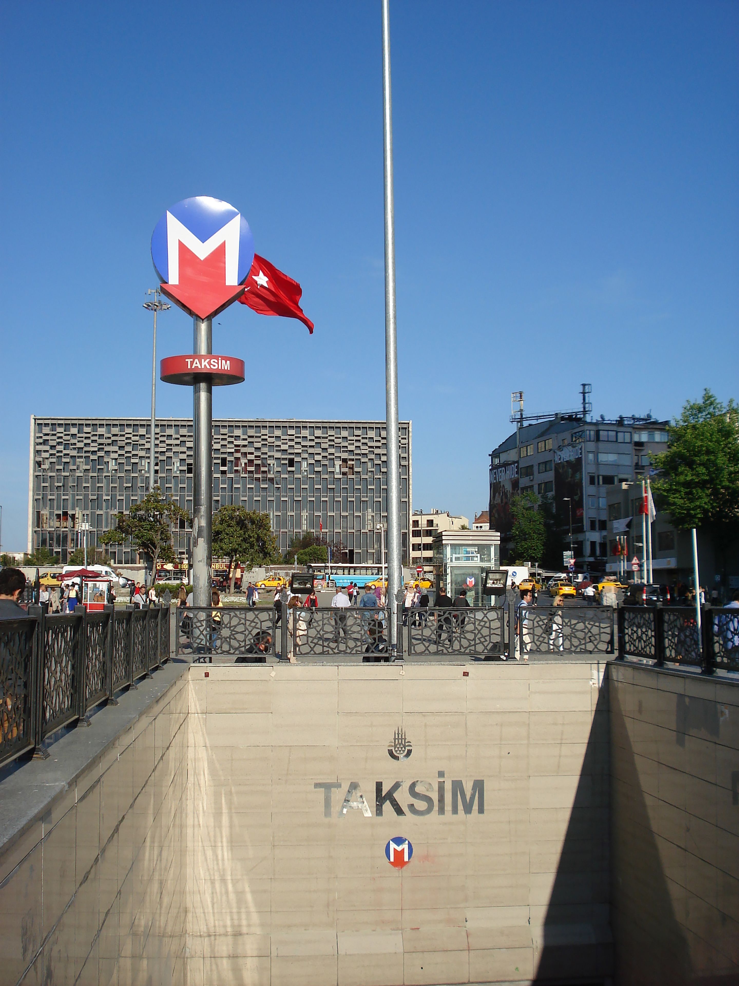 A public square with a prominent TAKSIM sign, a tall flagpole with a red flag, and a modern building in the background.