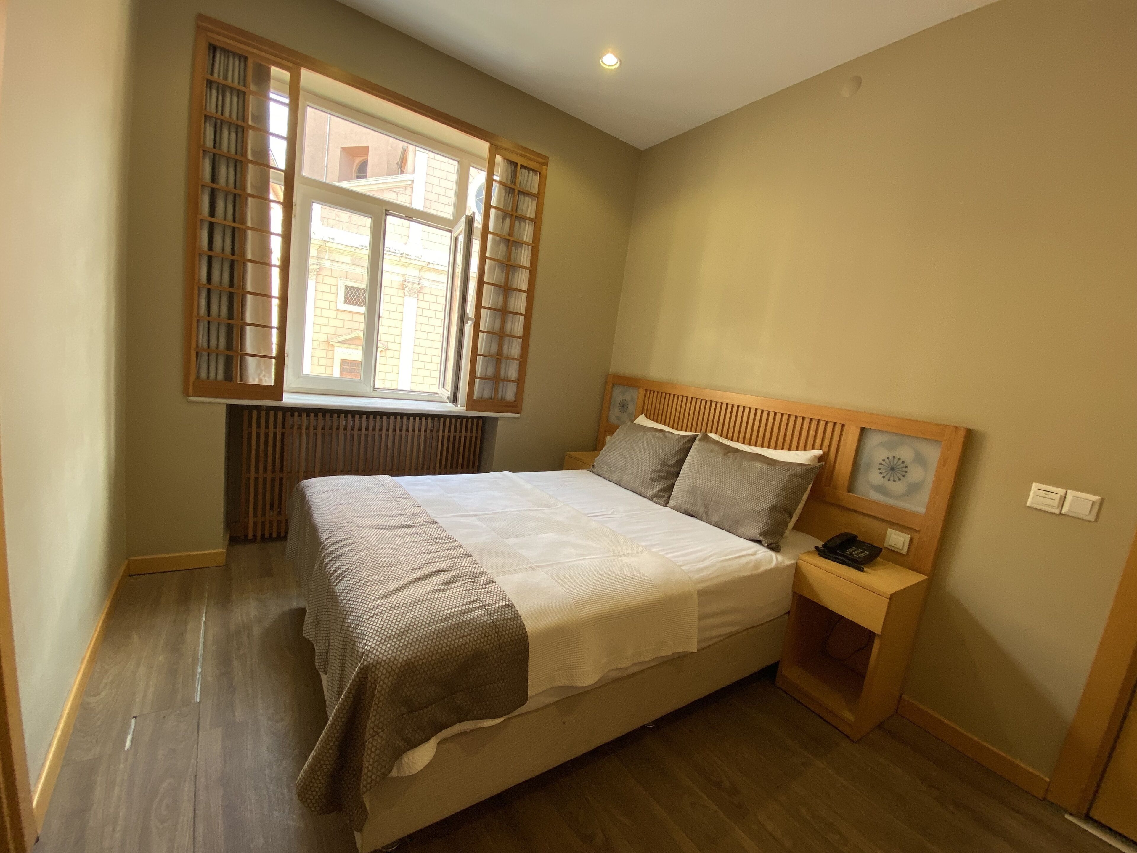 A bedroom with a bed, a bedside table, a nightstand, a window with blinds, and a radiator.