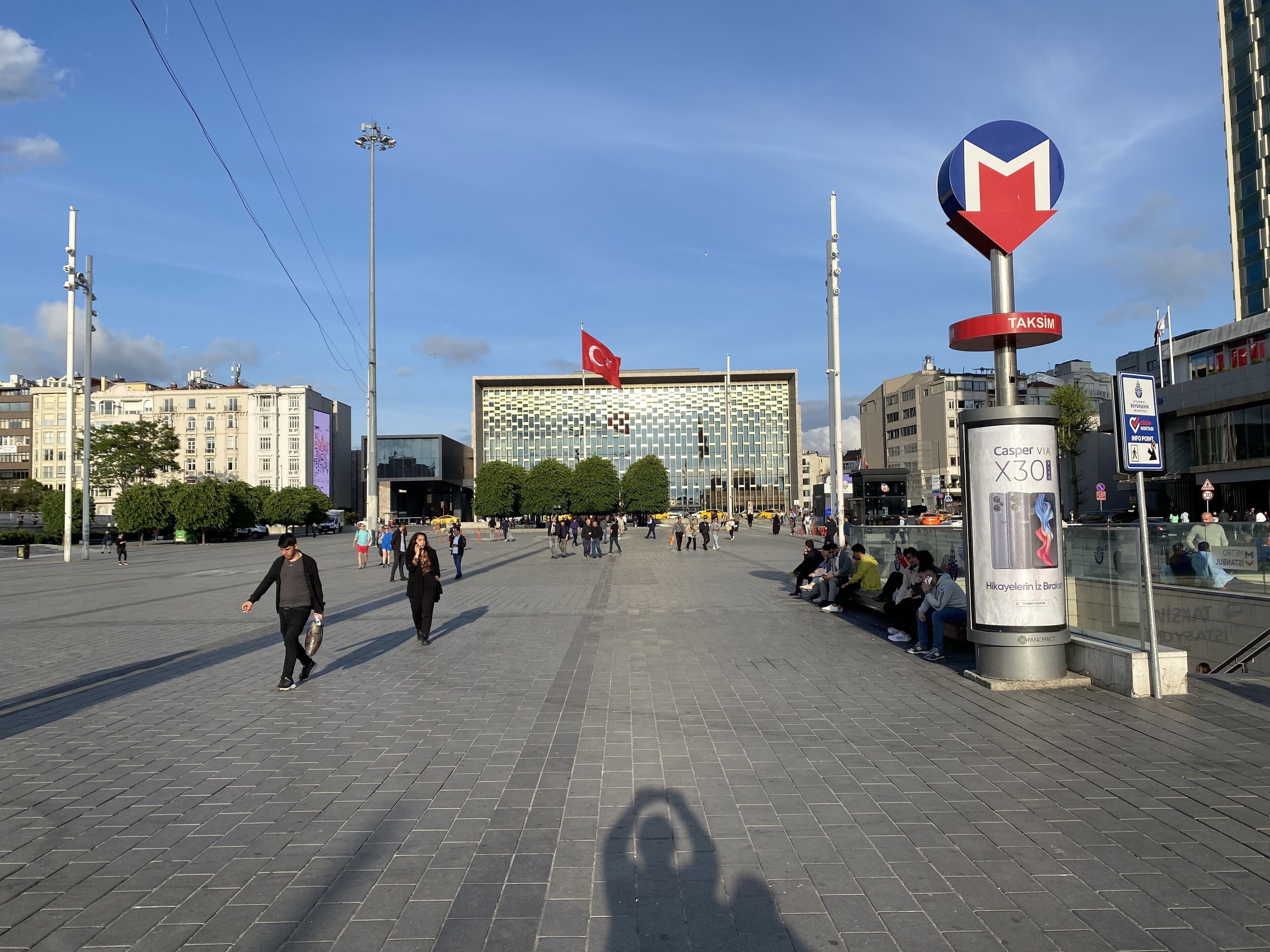 A public square with a modern building featuring a patterned facade, a flag, and a signboard advertising a mobile phone.