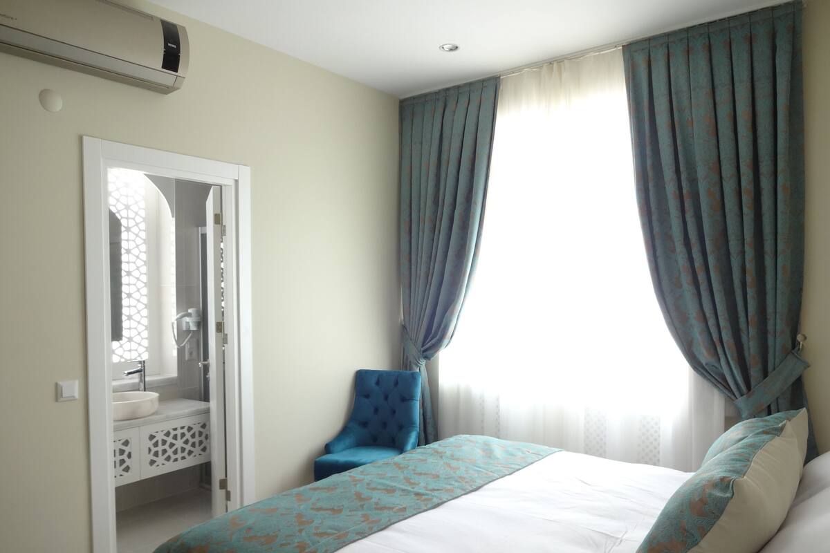 A bedroom with a bed, a chair, a bathroom with a sink, and a window with curtains.