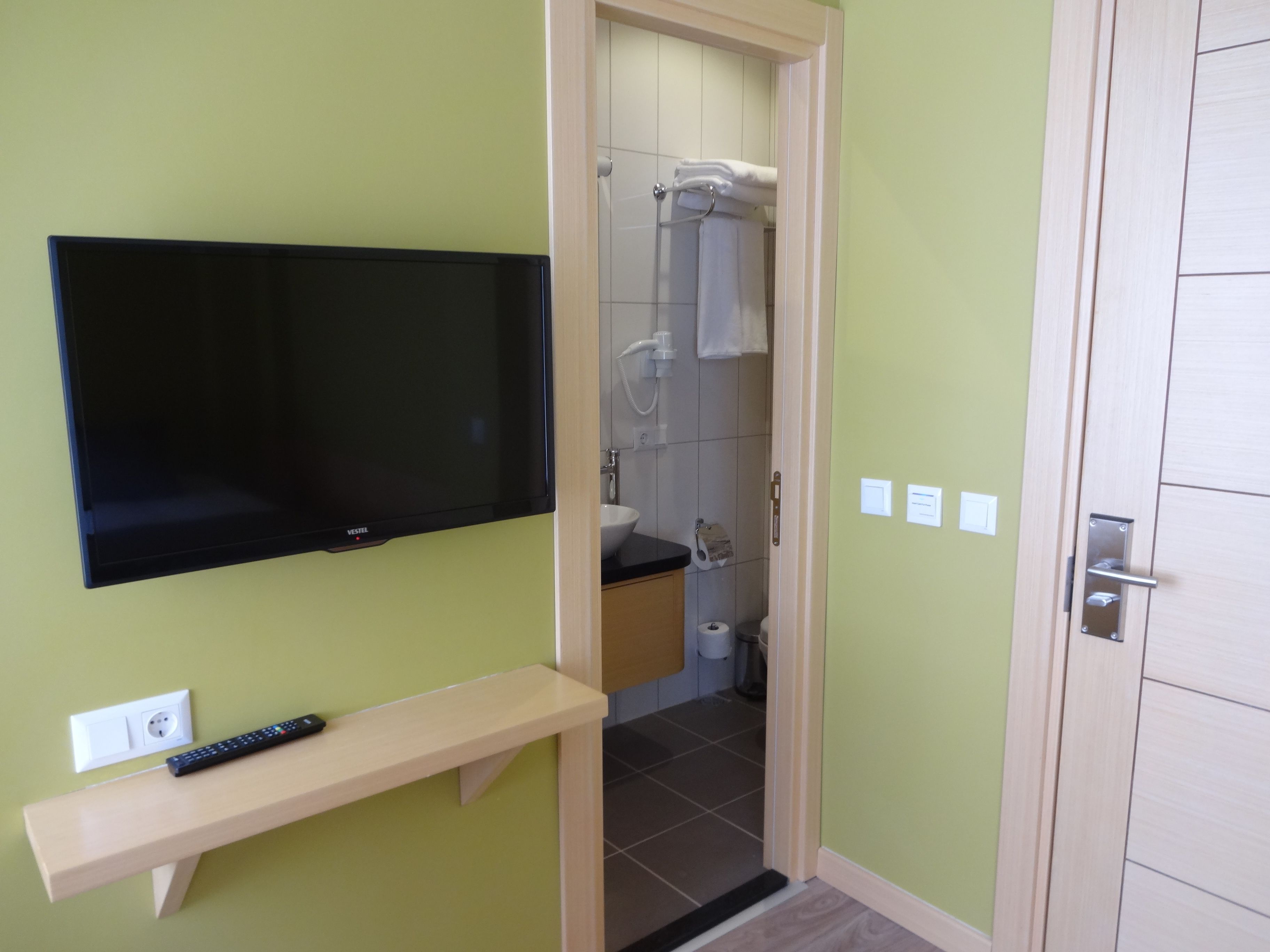 A bathroom with a shower, toilet, and sink. There is a TV mounted on the wall and a shelf with a remote control.