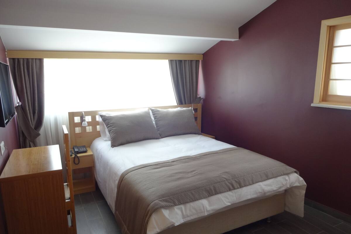 A bedroom with a bed, a wooden nightstand, a desk, and a window with curtains.