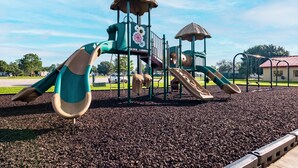 A playground with slides and climbing structures, surrounded by a gravel area and a clear sky.