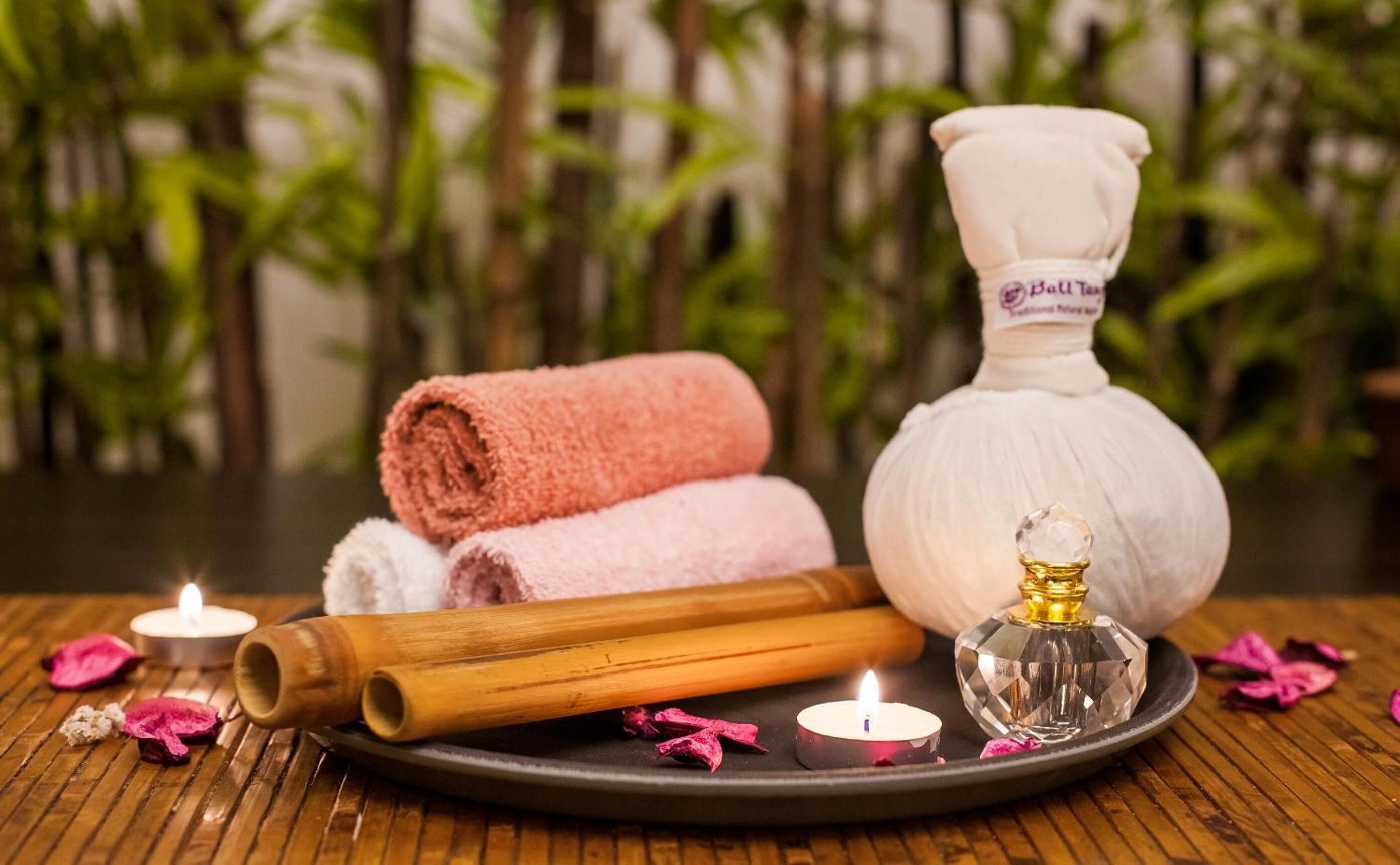 Wellness-Ambiente mit gerollten Handtüchern, einer Massageölflasche, einem Holzroller und Kerzen auf einem Tablett.