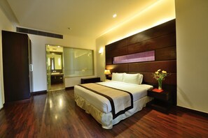 A modern hotel room with a large bed, a nightstand, a bedside table with a vase of flowers, and a large mirror. - Mapple Exotica (New Delhi)