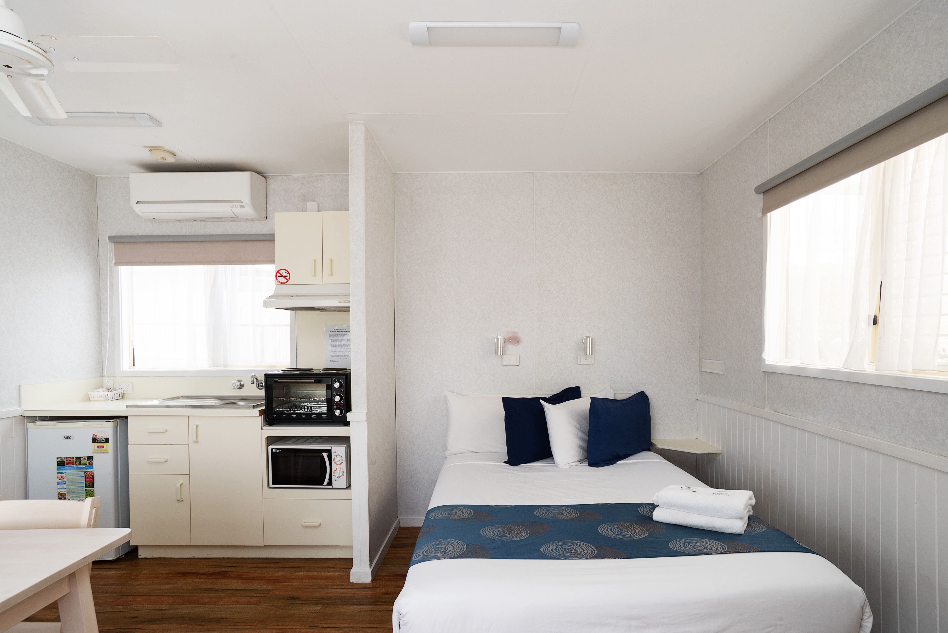 A compact hotel room with a kitchenette, a bed with blue and white bedding, and a window with curtains.