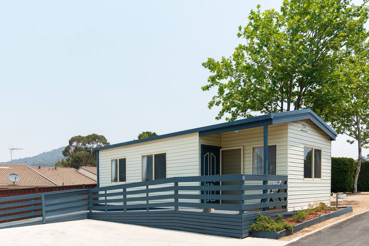A small, single-story mobile home with a blue roof and white siding, located in a residential area with a clear sky and a tree to the right.