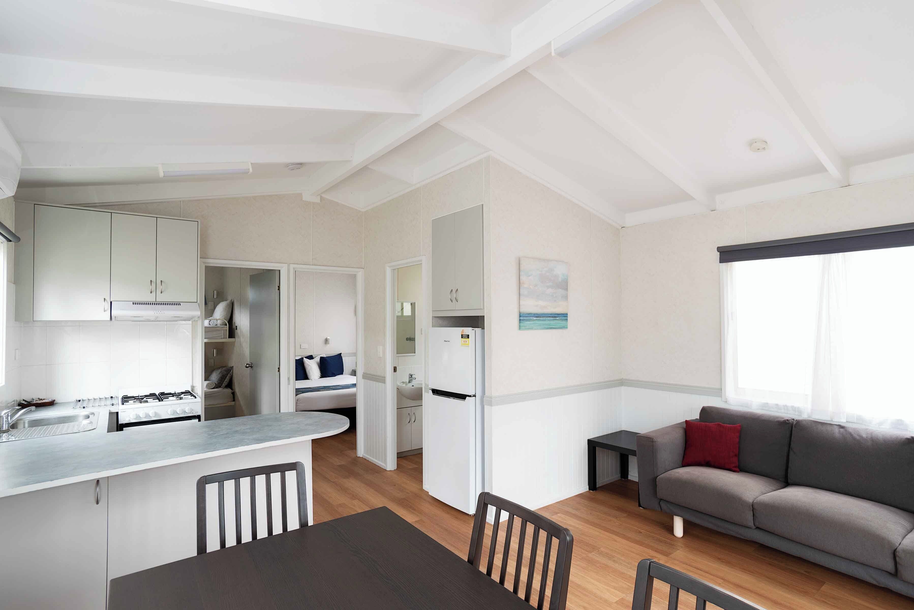 A modern kitchen with white cabinets, a dining area with a table and chairs, a gray sofa, and a window with sheer curtains.