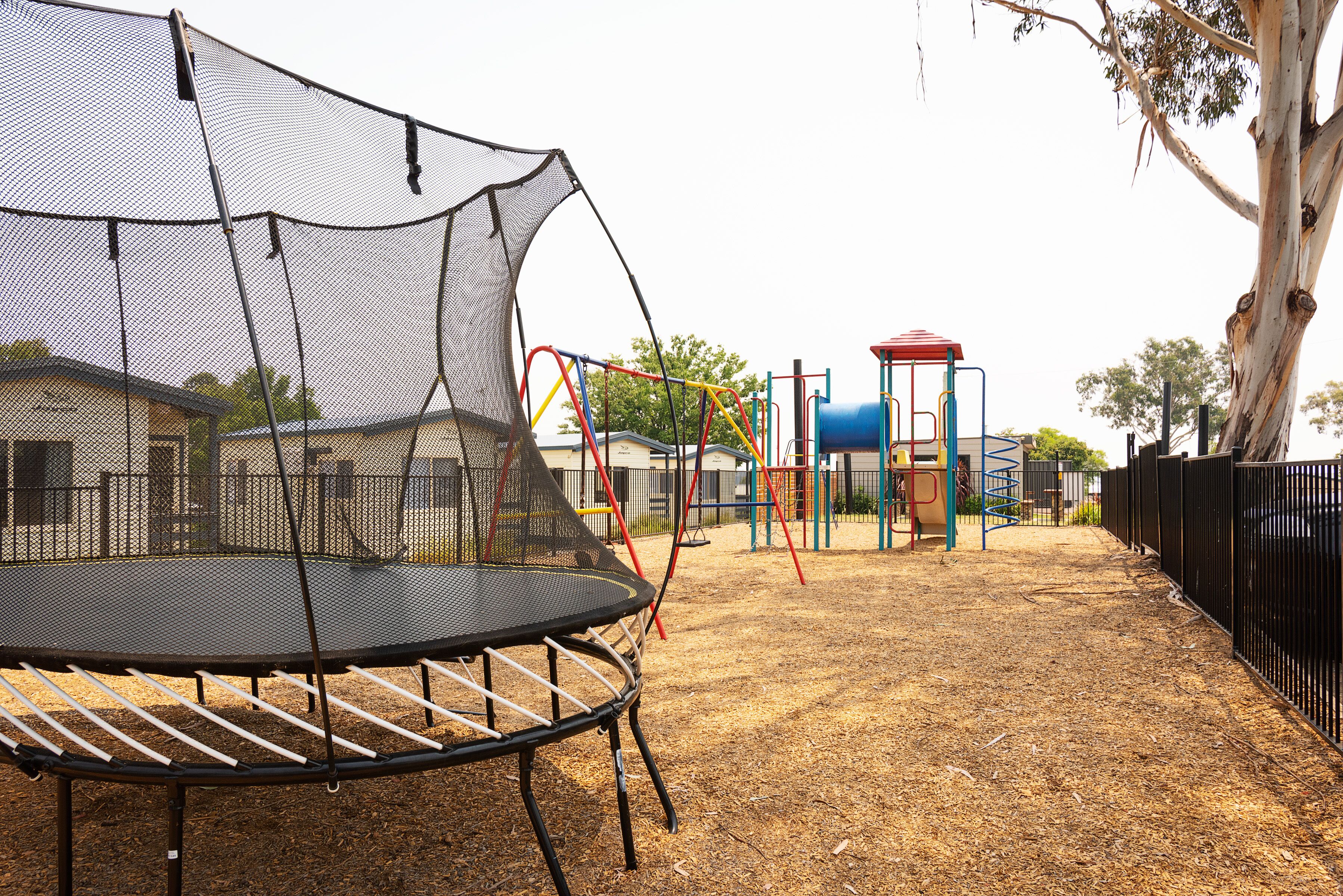 Un trampoline doté d’un filet de sécurité, une aire de jeux avec différentes structures ludiques et un espace délimité par une clôture.