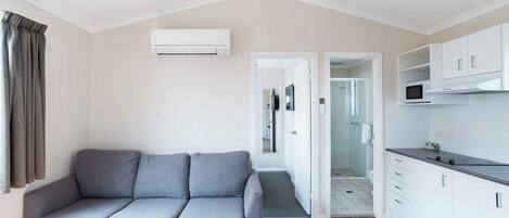 Una sala de estar moderna con un sofá gris, un aire acondicionado blanco y una cocina con armarios blancos y un microondas.