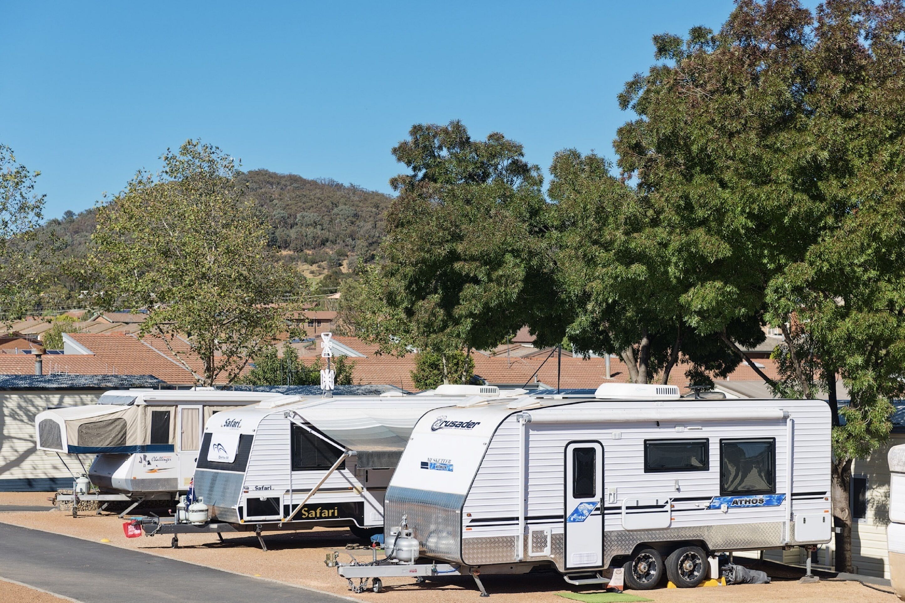 Remolques estacionados en un camping, con árboles y casas al fondo.