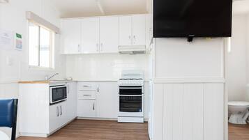A compact kitchen with white cabinets, a microwave, an oven, a sink, and a flat-screen TV mounted on the wall.