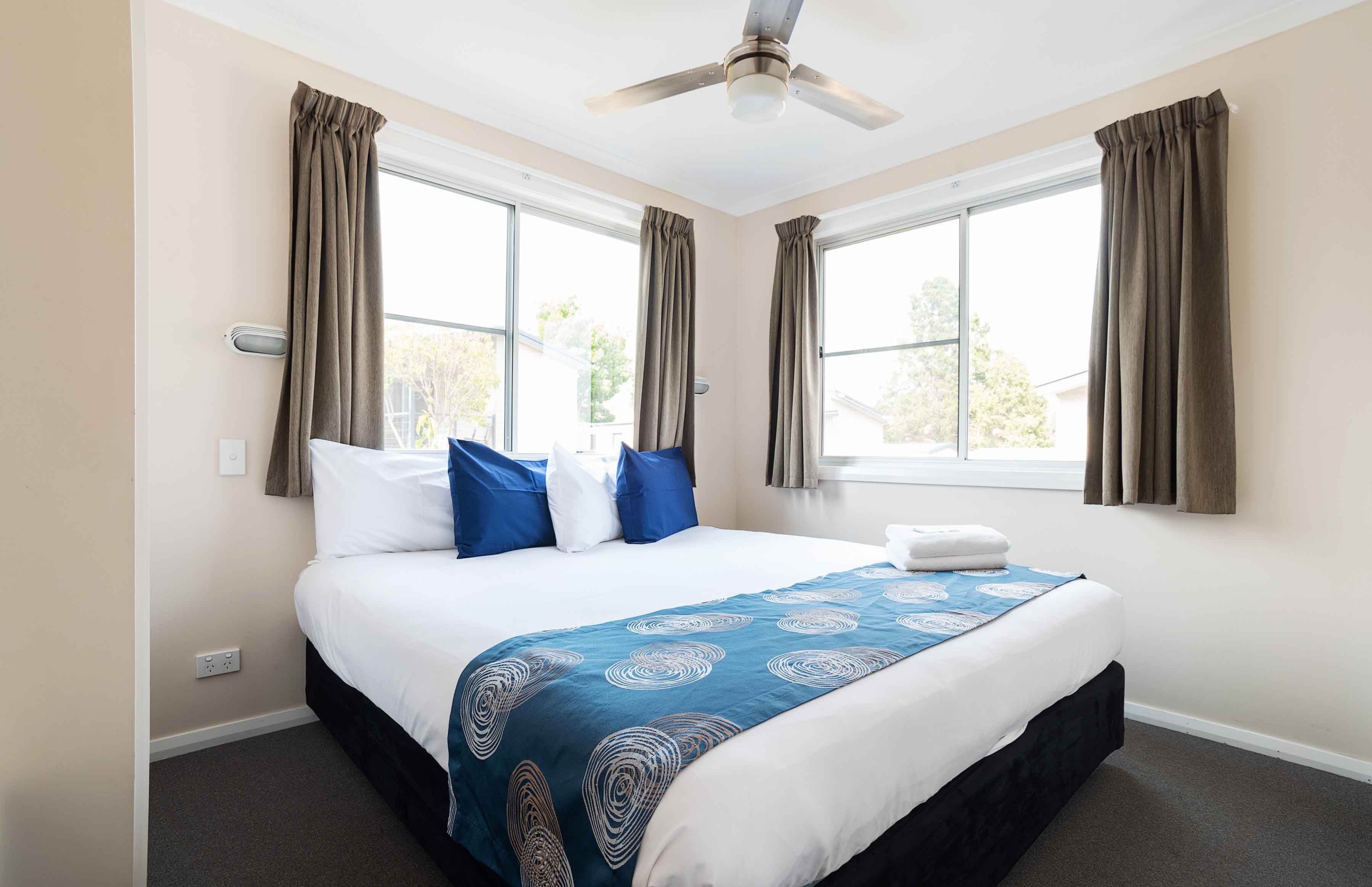 Una habitación de hotel con una cama, un ventilador de techo y dos ventanas con cortinas.