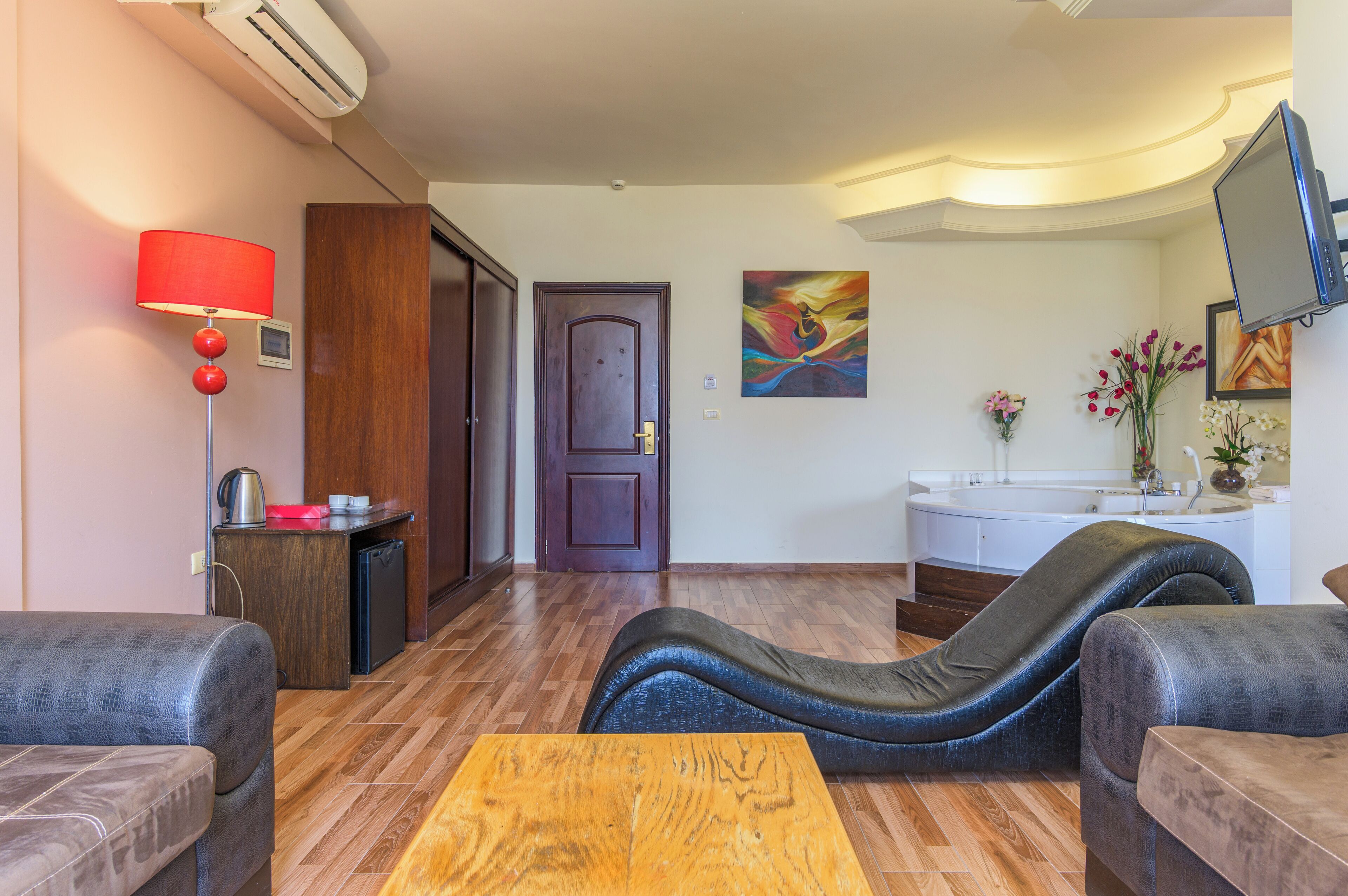 Una habitación con sofá, mesa de centro de madera, lámpara roja, cuadro, televisor y baño con lavamanos y flores.