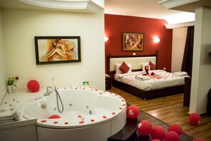 Habitación de hotel con una bañera grande, una cama con pétalos de rosa y una almohada con forma de corazón, y dos cuadros en las paredes.