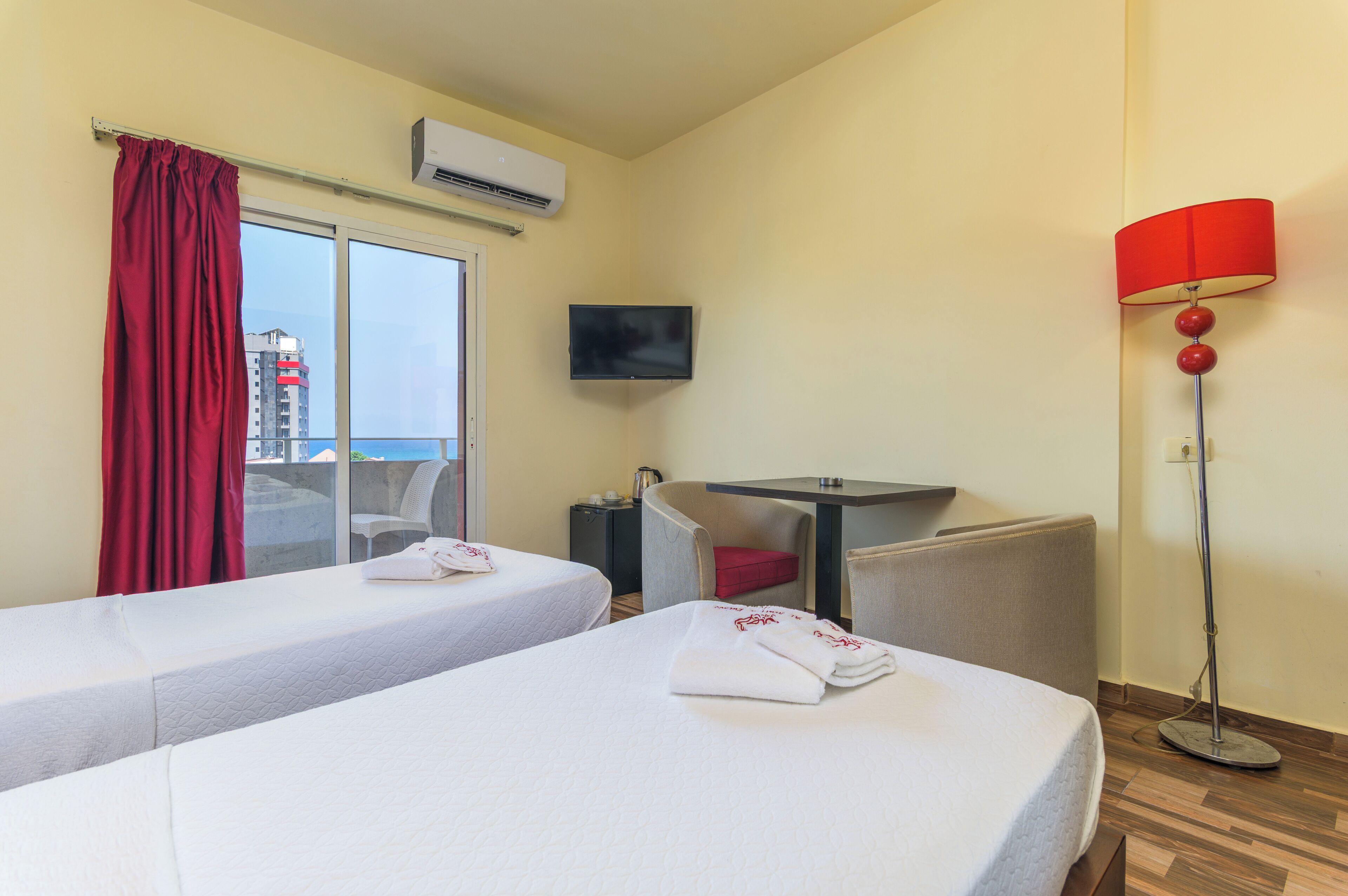 Habitación de hotel con dos camas, televisión, aire acondicionado, escritorio, silla, lámpara roja y vista a un edificio.