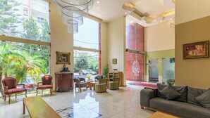 A spacious lobby with a large sofa, wooden tables, and a red patterned chair. There is a large window with a view of palm trees and a parked car outside. - Al Murjan Palace Hotel (Jounieh)