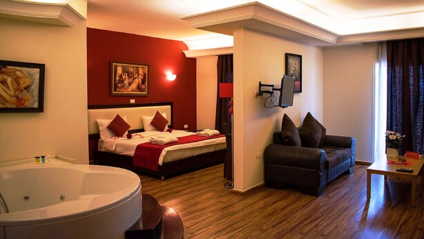 A hotel room with a large bed, a sofa, a coffee table, a TV, and a bathroom with a bathtub. - Al Murjan Palace Hotel (Jounieh)