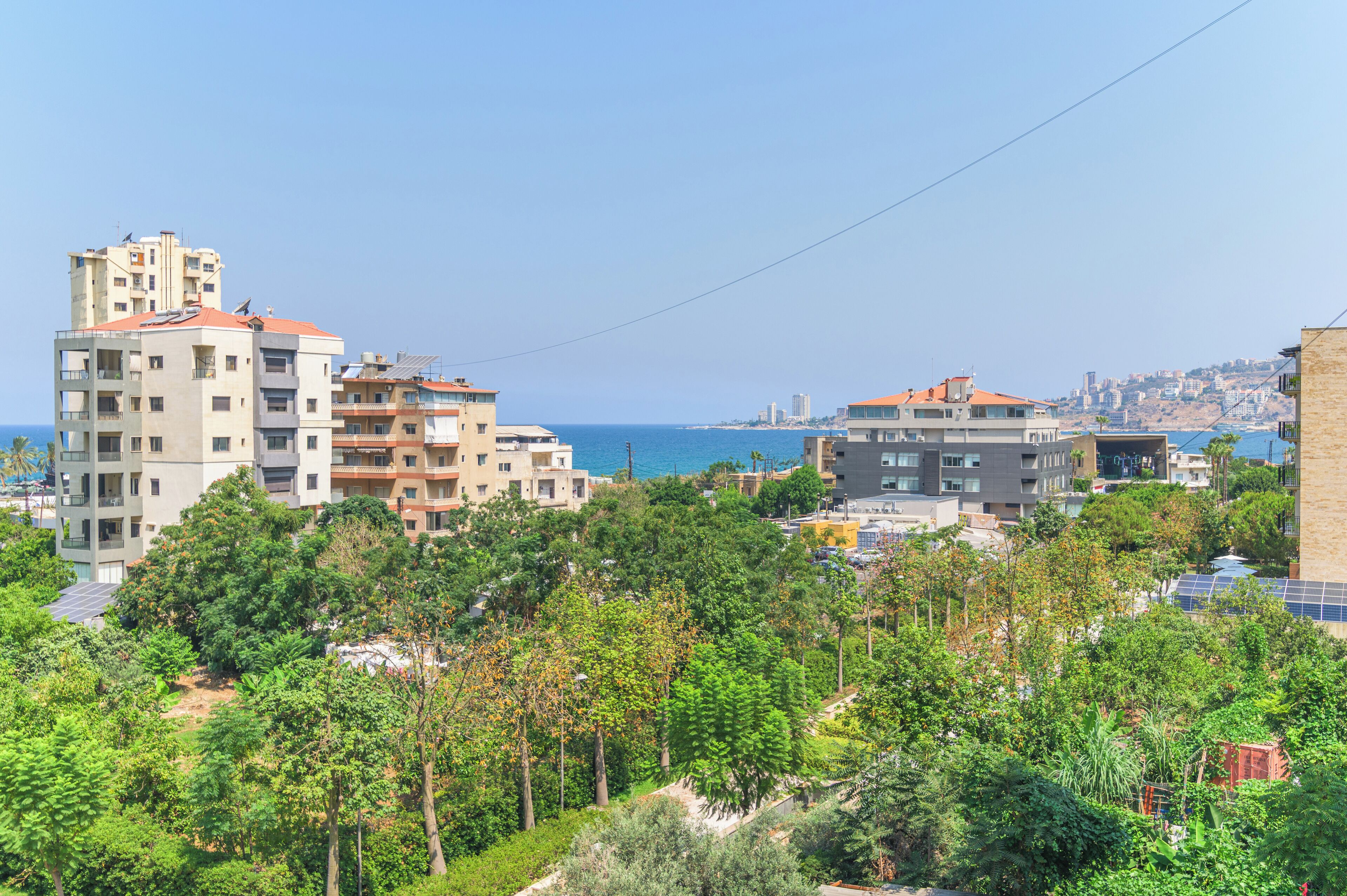 A coastal residential area with multi-story buildings, dense greenery, and a clear view of the sea.