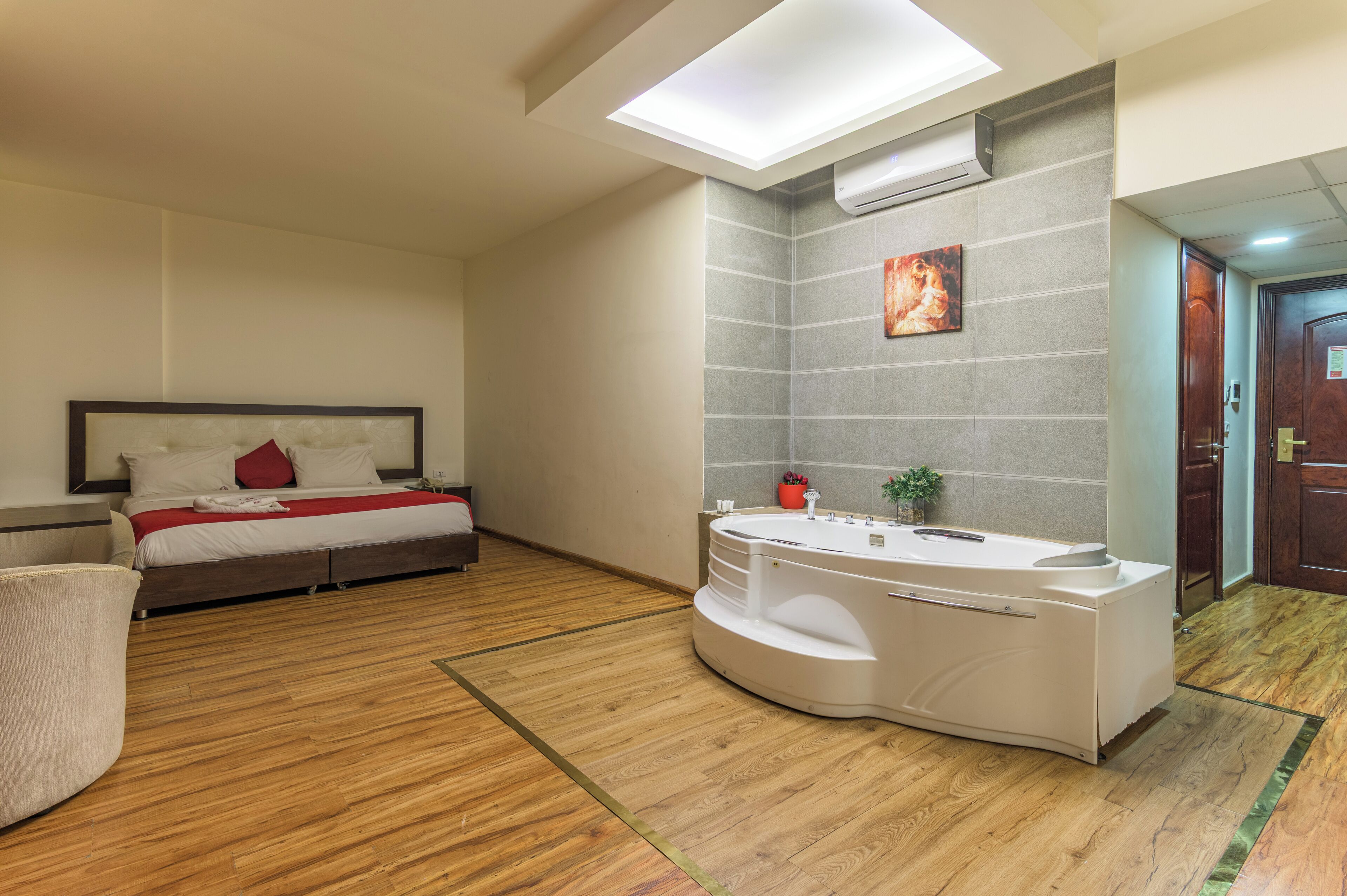 A modern hotel room with a bed, a large window, a built-in bathtub, and a wooden floor.