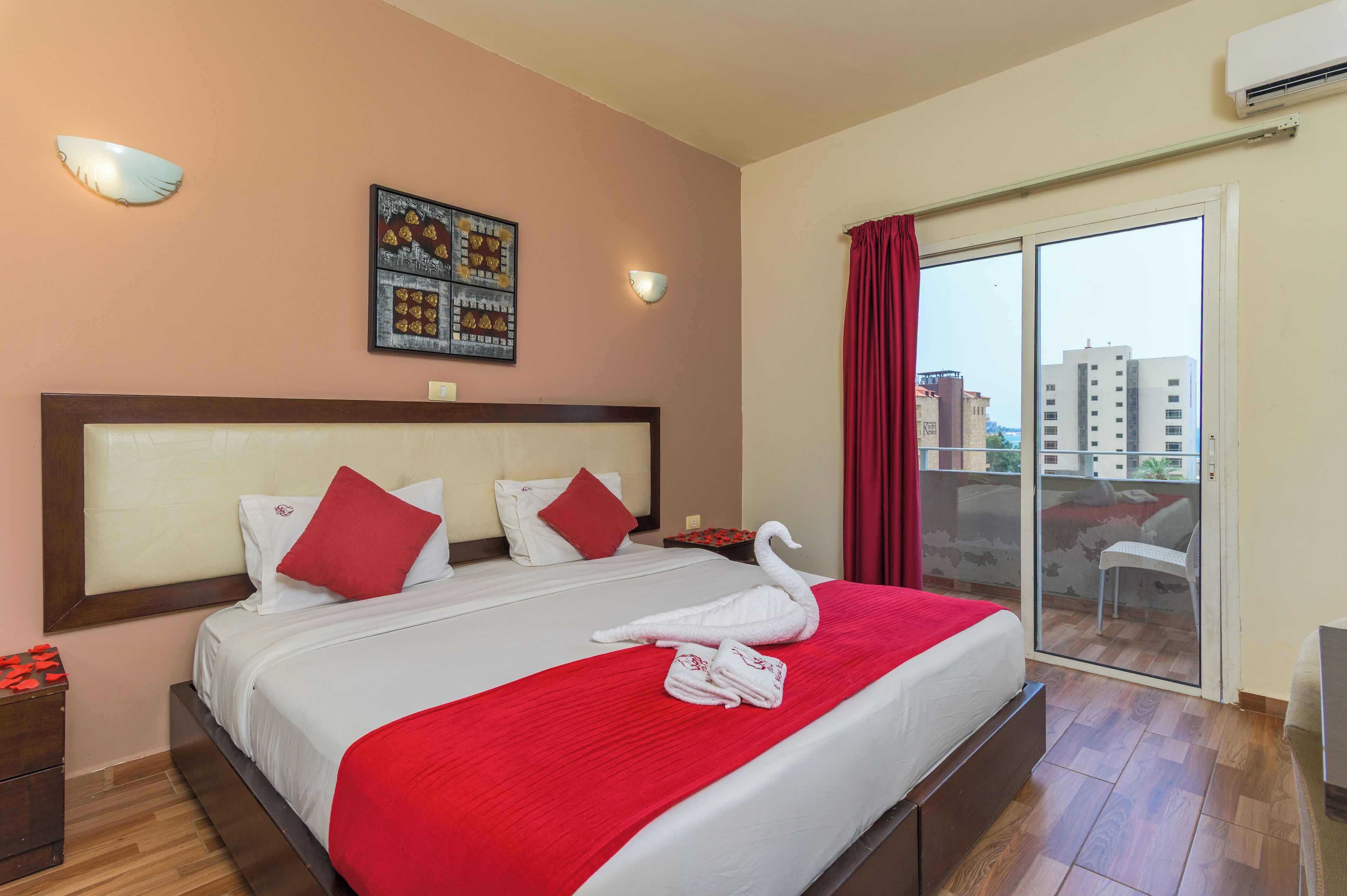 Habitación de hotel con una cama grande, decoración en colores rojo y blanco, un balcón con vistas a edificios y un aire acondicionado de pared.