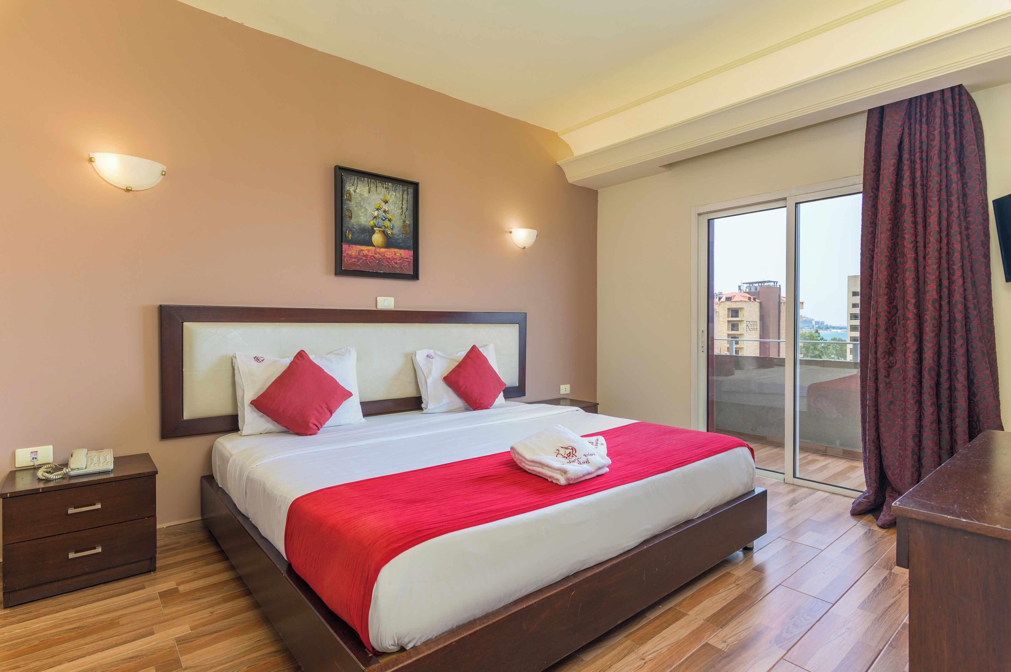 Un cuarto de hotel con una cama grande, una mesita de noche, un cuadro en la pared y vistas a través de un ventanal corredero.