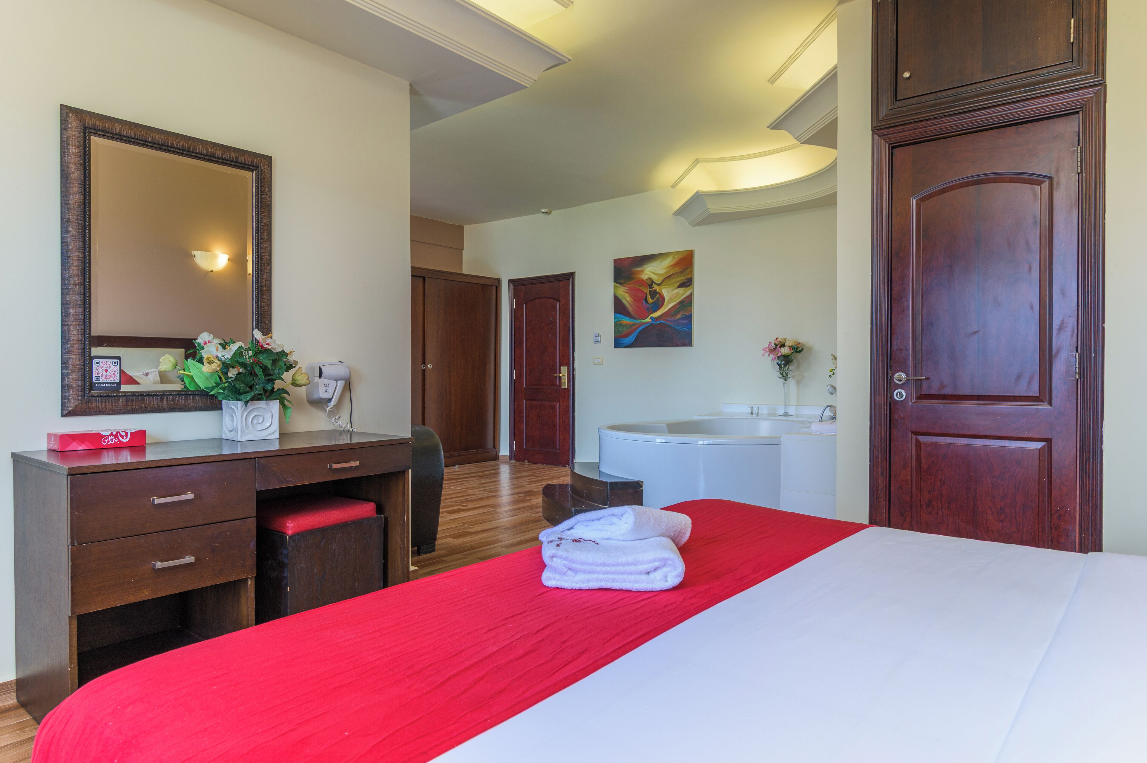 Hotelkamer met een bed, een bureau met lades, een spiegel, een schilderij, een badkamer met een wastafel en spiegel, en een deur.