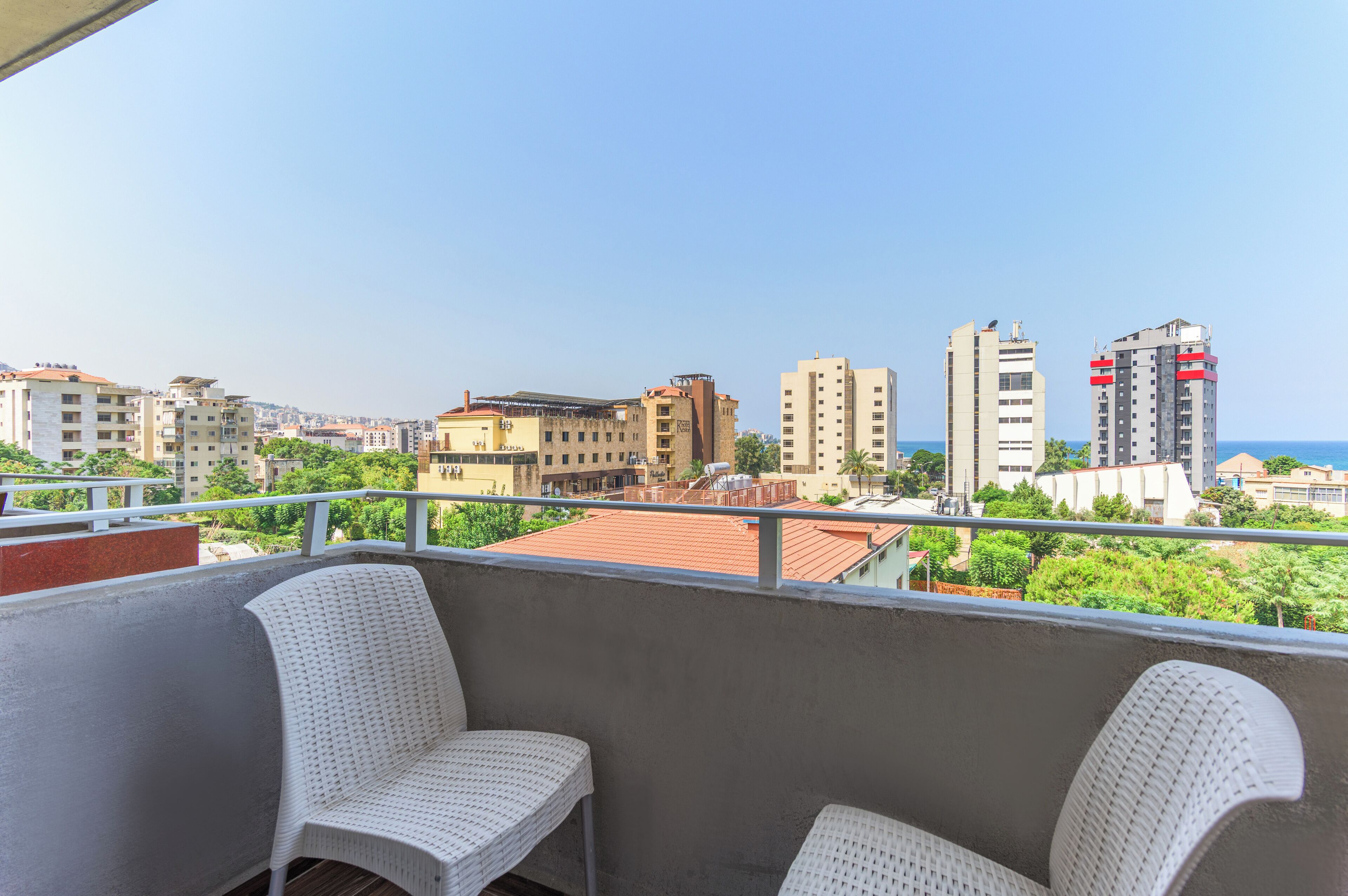 Ein Balkon mit zwei weißen Korbstühlen, Blick auf eine Stadtlandschaft mit verschiedenen Gebäuden und einem klaren Himmel.