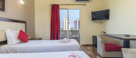 Habitación de hotel con dos camas, un televisor, un escritorio y vistas a edificios a través de una ventana.